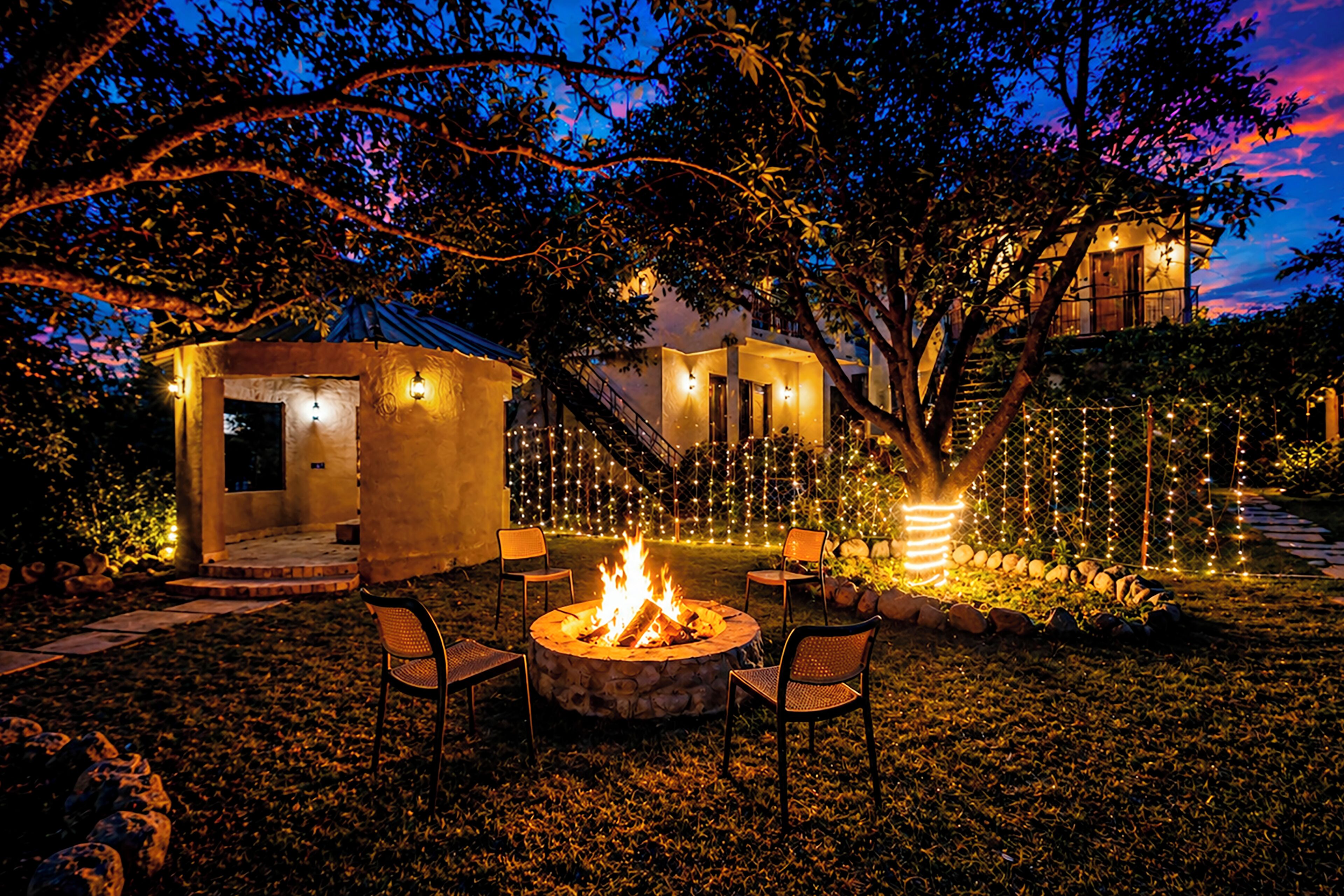 A fire pit with chairs, a stone structure, and string lights in a garden at dusk.