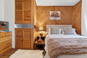 Ein Schlafzimmer mit einem hölzernen Kleiderschrank, einem Nachttisch mit Lampe, einem Bett mit vielen Kissen und einem Wandbild.