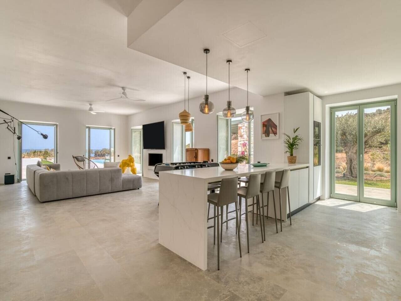A spacious living area with a large sofa, a kitchen island with bar stools, and large windows offering views of the outdoors.
