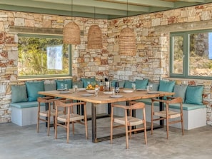 Ein Essbereich mit Steinwänden, einem Holztisch und -stühlen, einer Steinsitzbank mit Kissen sowie gewebten Pendelleuchten.