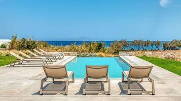 A pool with three lounge chairs facing the water, surrounded by a patio area with a clear view of the sea and distant mountains.