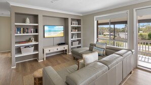 A living room with a sofa, TV, bookshelf, and a balcony with a view. - Ocean Vibes - Your Ultimate Scotts Head Coastal Escape (Scotts Head)