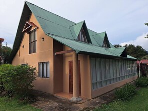 A two-story house with a green metal roof, a large window with curtains, and a small porch area. - Casa Luna is Your Best Option Near Poás Volcano! (Sabana Redonda)