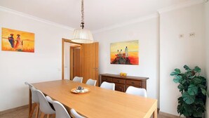 Un comedor con una mesa de madera, sillas blancas, una araña y dos cuadros en las paredes.
