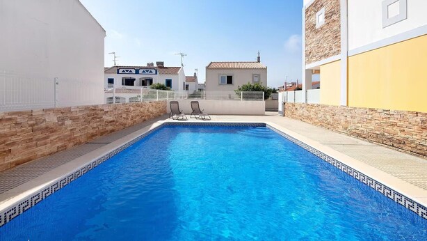 Una piscina con dos sillas de descanso, rodeada de muros de piedra y edificios.