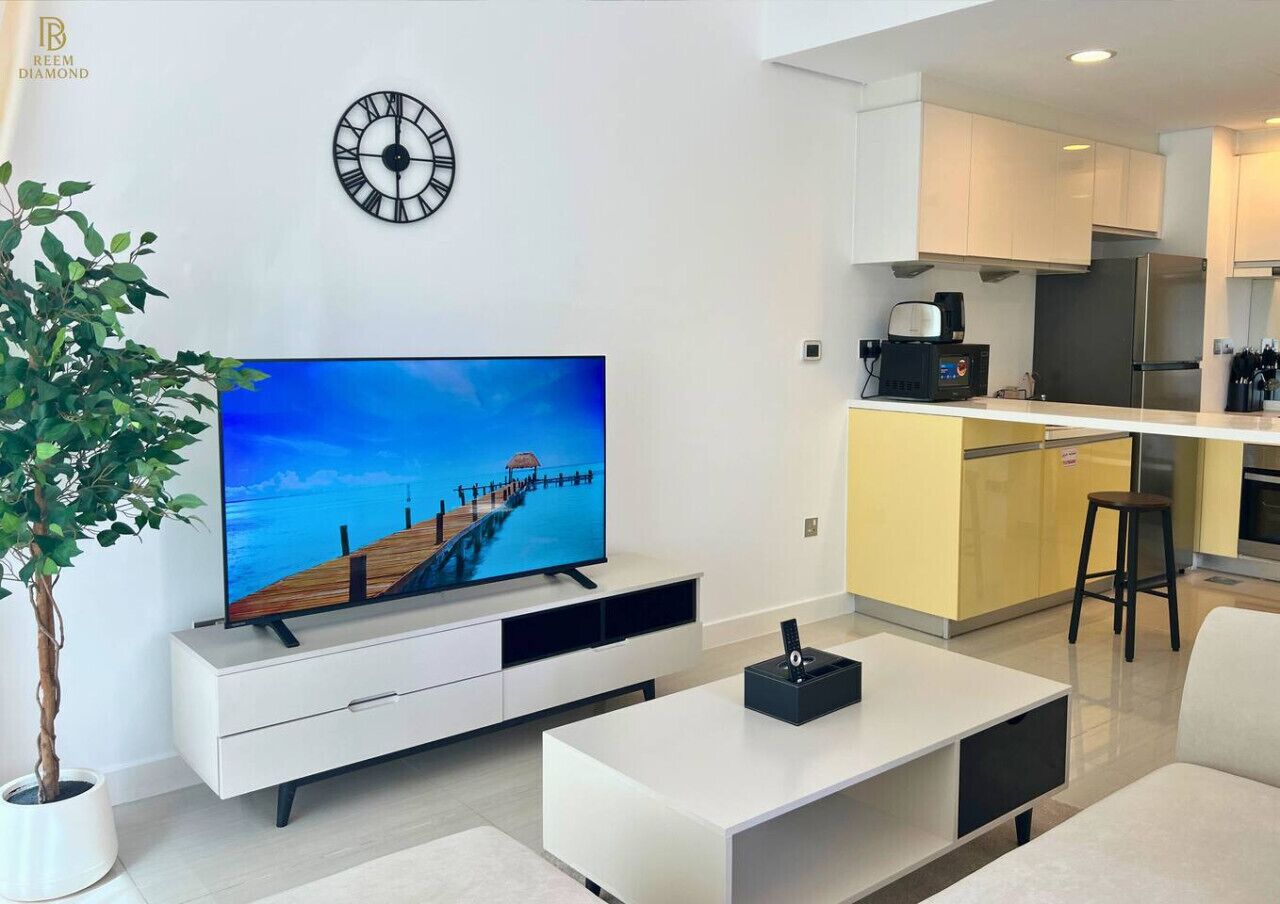 A modern living room with a flat-screen TV, a clock, a plant, and a kitchen area.