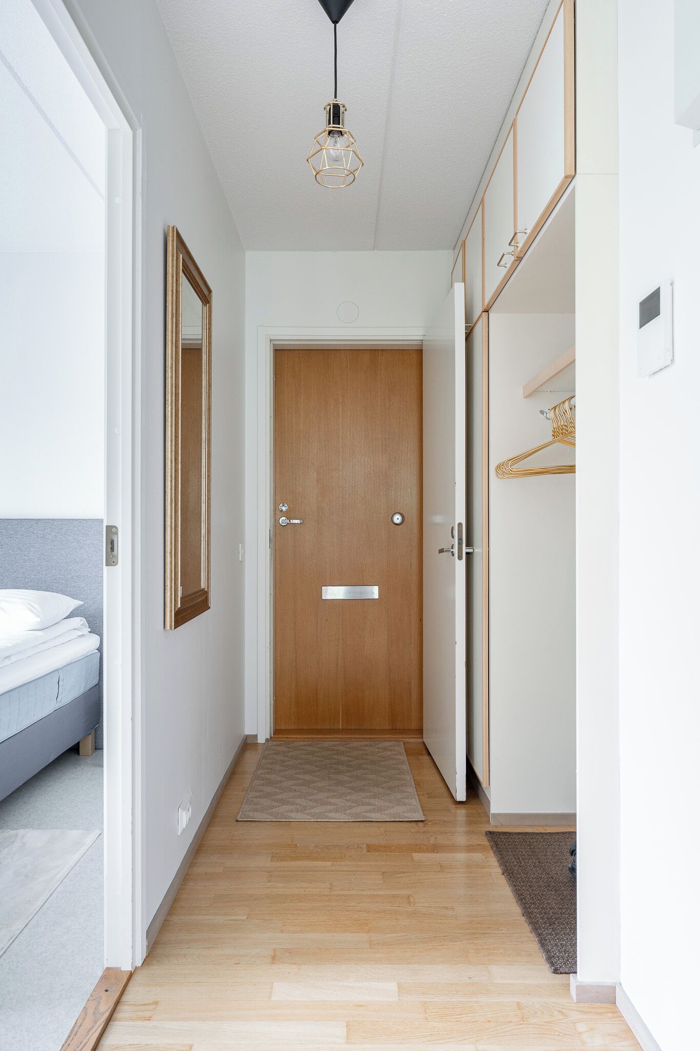 Een hal met een houten deur, een bed met een witte matras, een hangend spiegel en een lamp.