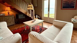 En stue med trappe, en hvid sofa, et lille bord og havudsigt gennem en dør.