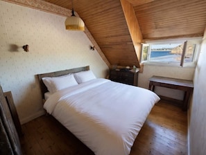 Ein Schlafzimmer mit Holzbalkendecke, einem Bett mit weißer Bettwäsche, einem Nachttisch, einer Kommode und einem Fenster mit Meerblick.