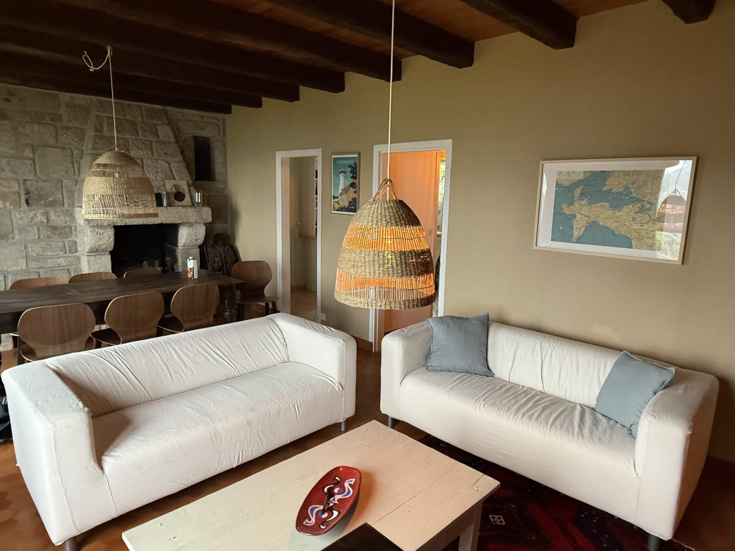 A living room with two white sofas, a wooden coffee table, a hanging woven light fixture, a stone fireplace, and a framed map on the wall.