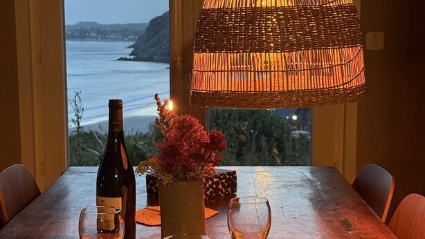 Et spisebord med en vinflaske, to vinglas, en vase med blomster og en vævet lampe.