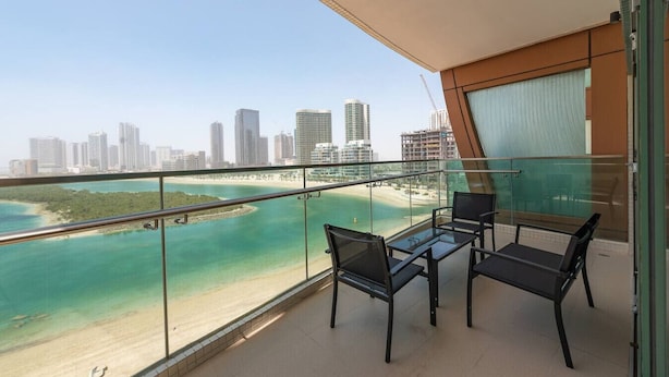 Un balcone con ringhiera in vetro, due sedie, un tavolino e vista su spiaggia e skyline della città.