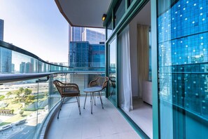 Un balcón con baranda de vidrio, dos sillas de mimbre, una mesita y vistas a un paisaje urbano con edificios altos.