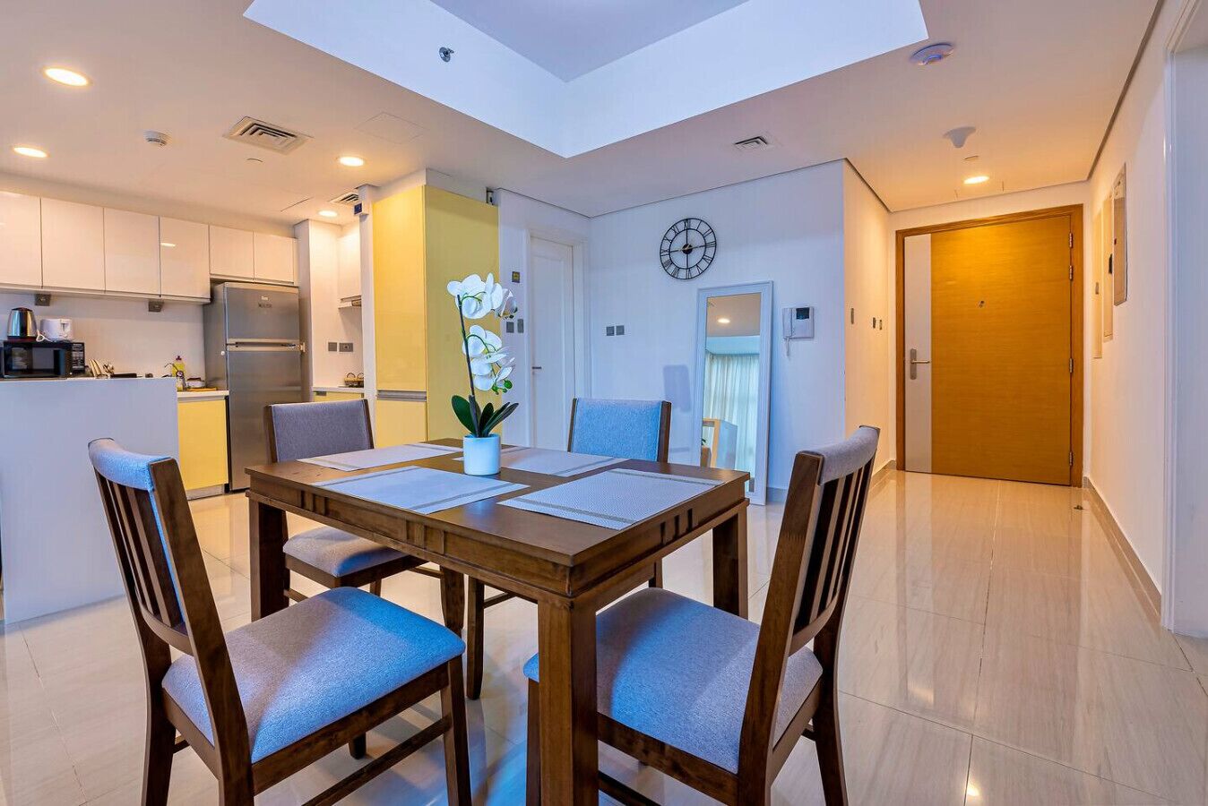 A modern kitchen with a dining area, a clock on the wall, and a door leading to another room.