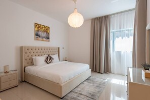 A bedroom with a tufted headboard, a bed with white linens, a bedside table with a lamp, a large window with curtains, and a painting on the wall. - Lavish 2BR Apt next to beach (Abu Dhabi)