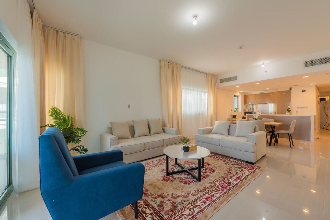 A modern living room with two sofas, a blue armchair, a coffee table, and a dining area with a table and chairs.