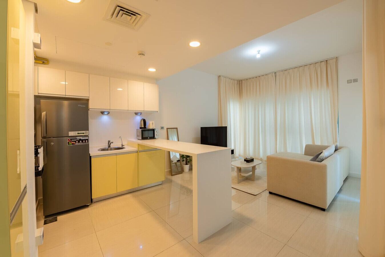 A modern kitchen with a built-in refrigerator, a microwave, and a sink. There is a small island with a table and chairs, and a sofa in the living area.