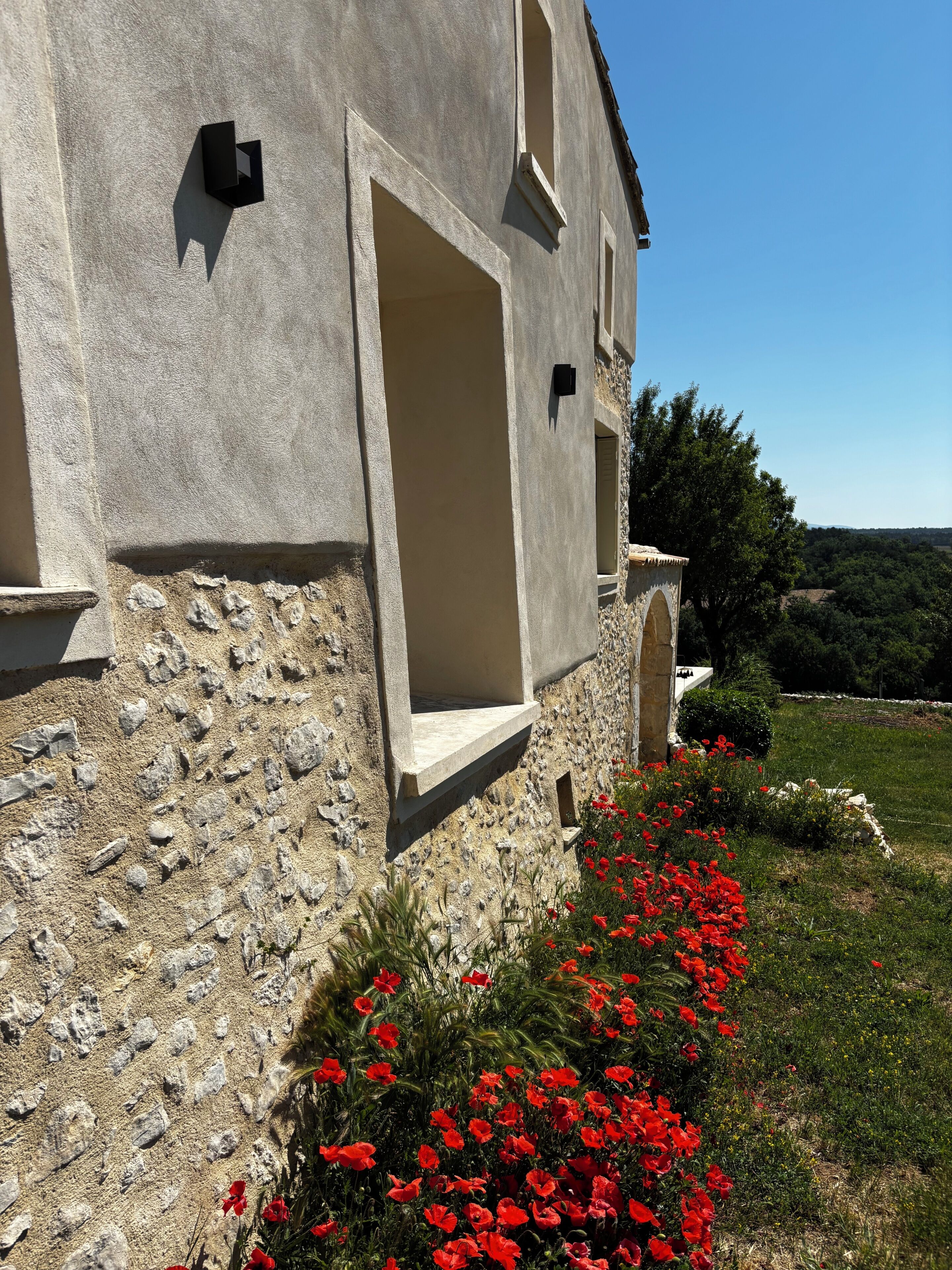 Un bâtiment en pierre doté d’une grande porte ouverte, d’une petite fenêtre et d’un parterre fleuri où poussent des fleurs rouges.