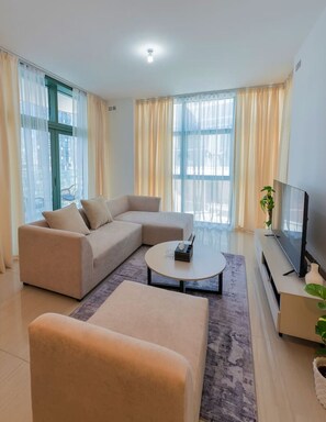 A modern living room with a sectional sofa, a round coffee table, a flat-screen TV, and large windows with sheer curtains. - Stylish & Cozy 1BR Apt in Reem Island (Abu Dhabi)
