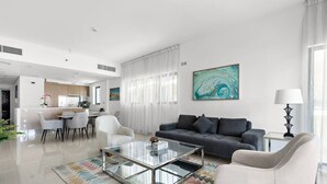 Un salon moderne avec un canapé, une table basse et des fauteuils. On aperçoit à l’arrière-plan un coin cuisine avec une table à manger et des chaises.