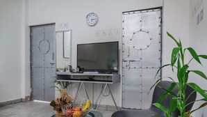 A room with a television, two metal doors, a clock, a glass table with fruits, and a chair.