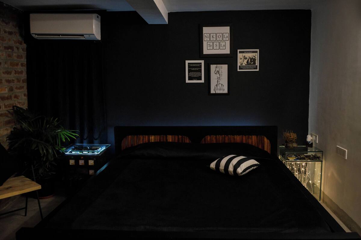 A dark-themed bedroom with a bed, a wall-mounted air conditioner, and framed artwork.