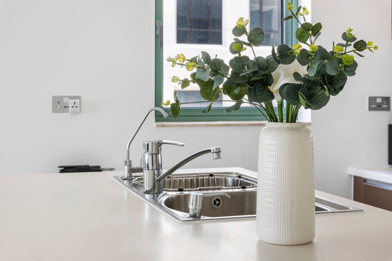 Una cocina moderna con un lavamanos de acero inoxidable, un jarrón blanco con follaje verde y una ventana con vistas al exterior.
