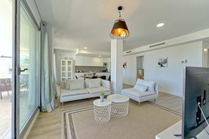 A modern living room with a sofa, two round coffee tables, a TV, and a kitchen in the background. - 3015 Luxury Serenity Views Apartment in Estepona (Buenas Noches)