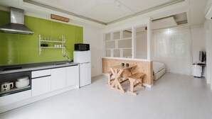 Eine kleine Küche mit grüner Rückwand, weißen Schränken, einem Holztisch und einem Bett im angrenzenden Zimmer.