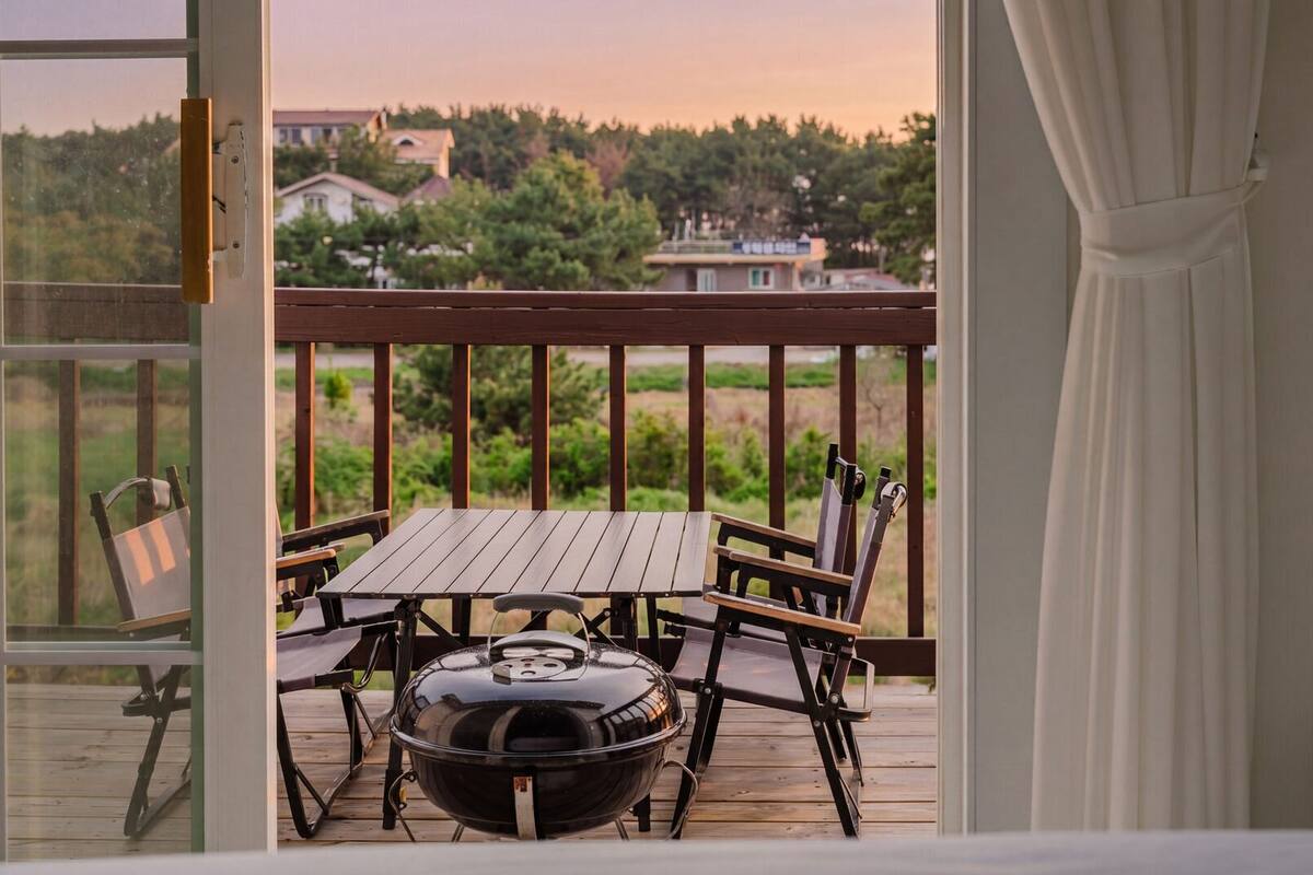 Una terraza de madera con una mesa, sillas, una parrilla y vistas a casas y árboles.