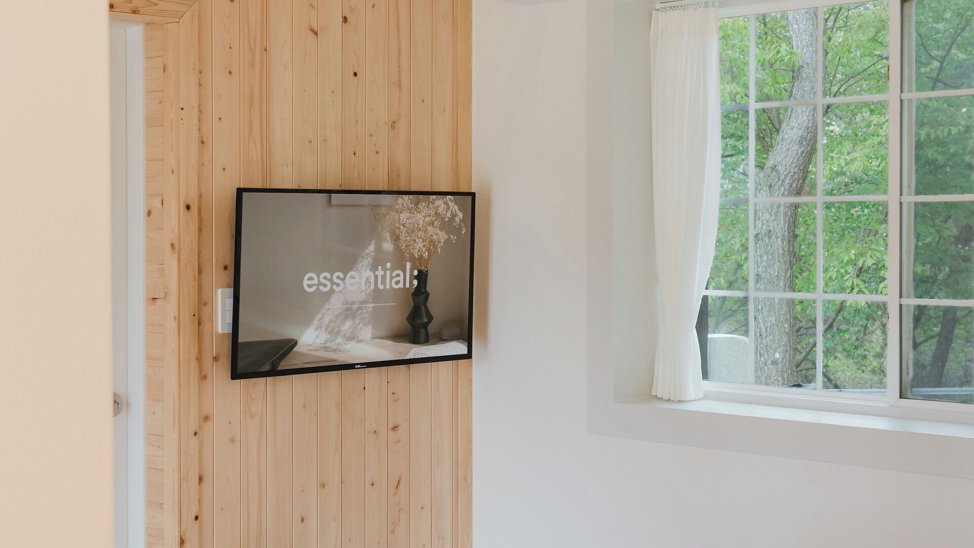 Un televisor montado en una pared de madera, una ventana con vista a árboles y una cortina blanca.