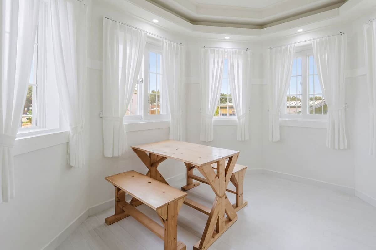 Una habitación con una mesa de madera y un banco, paredes blancas y varias ventanas con cortinas blancas.