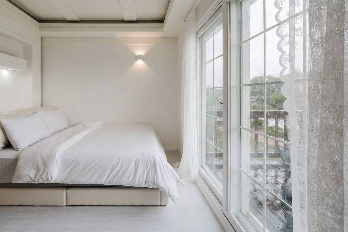Un dormitorio con una cama, una ventana con cortinas de encaje y una lámpara fijada a la pared.