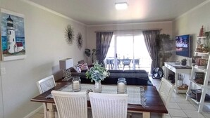 A dining area with a table and chairs, a painting of a lighthouse, a television, and a plant. - Secure 2-bedroom Apartment With Pool View in Mossel Bay (Mossel Bay)