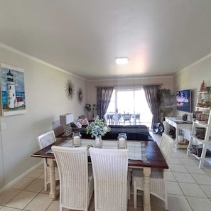 A dining area with a table and chairs, a painting of a lighthouse, a television, and a plant. - Secure 2-bedroom Apartment With Pool View in Mossel Bay (Mossel Bay)