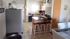A compact kitchen with a refrigerator, stove, and sink. There are stools at the counter and a dining table with chairs. - Secure 2-bedroom Apartment With Pool View in Mossel Bay (Mossel Bay)