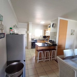 A compact kitchen with a refrigerator, stove, and sink. There are stools at the counter and a dining table with chairs. - Secure 2-bedroom Apartment With Pool View in Mossel Bay (Mossel Bay)