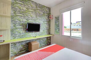 Una habitación con pared de piedra, un televisor montado en ella, una ventana con vistas a edificios y una cama con colcha roja y blanca.