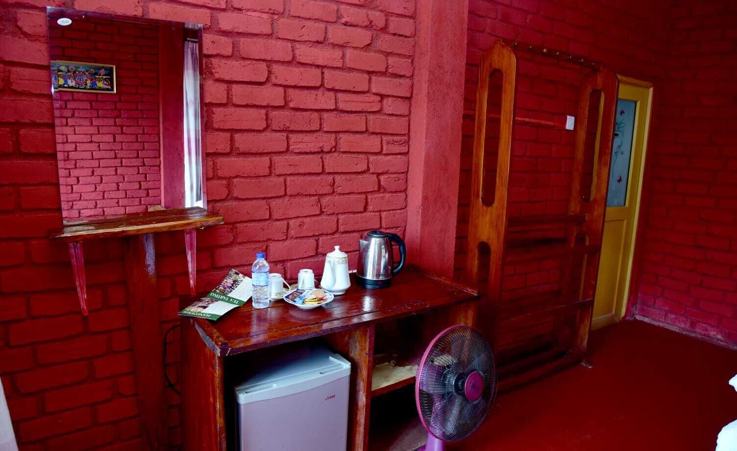 Une pièce aux murs de briques rouges, avec une table en bois, un ventilateur, une bouilloire et un petit réfrigérateur.