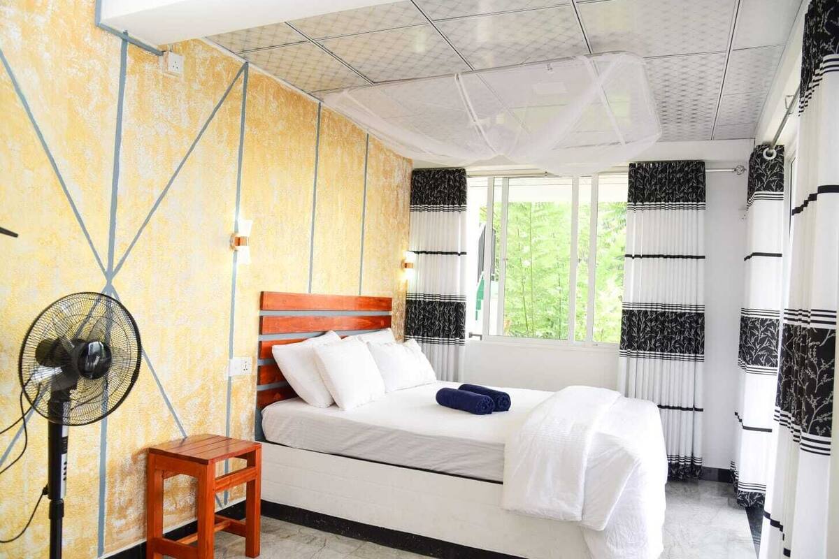 A bedroom with a bed, a standing fan, and a small wooden table.