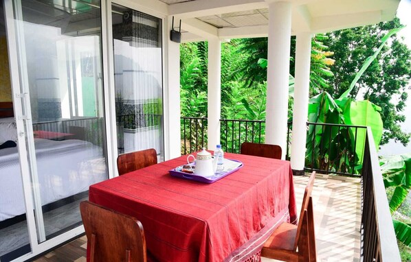 Ein Balkon mit gedecktem Frühstückstisch, einem Bett mit roter Bettdecke und üppigem Grün im Hintergrund.