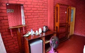 Una habitación con pared de ladrillo rojo, una mesa de madera con un ventilador, un refrigerador pequeño y un armario de madera.