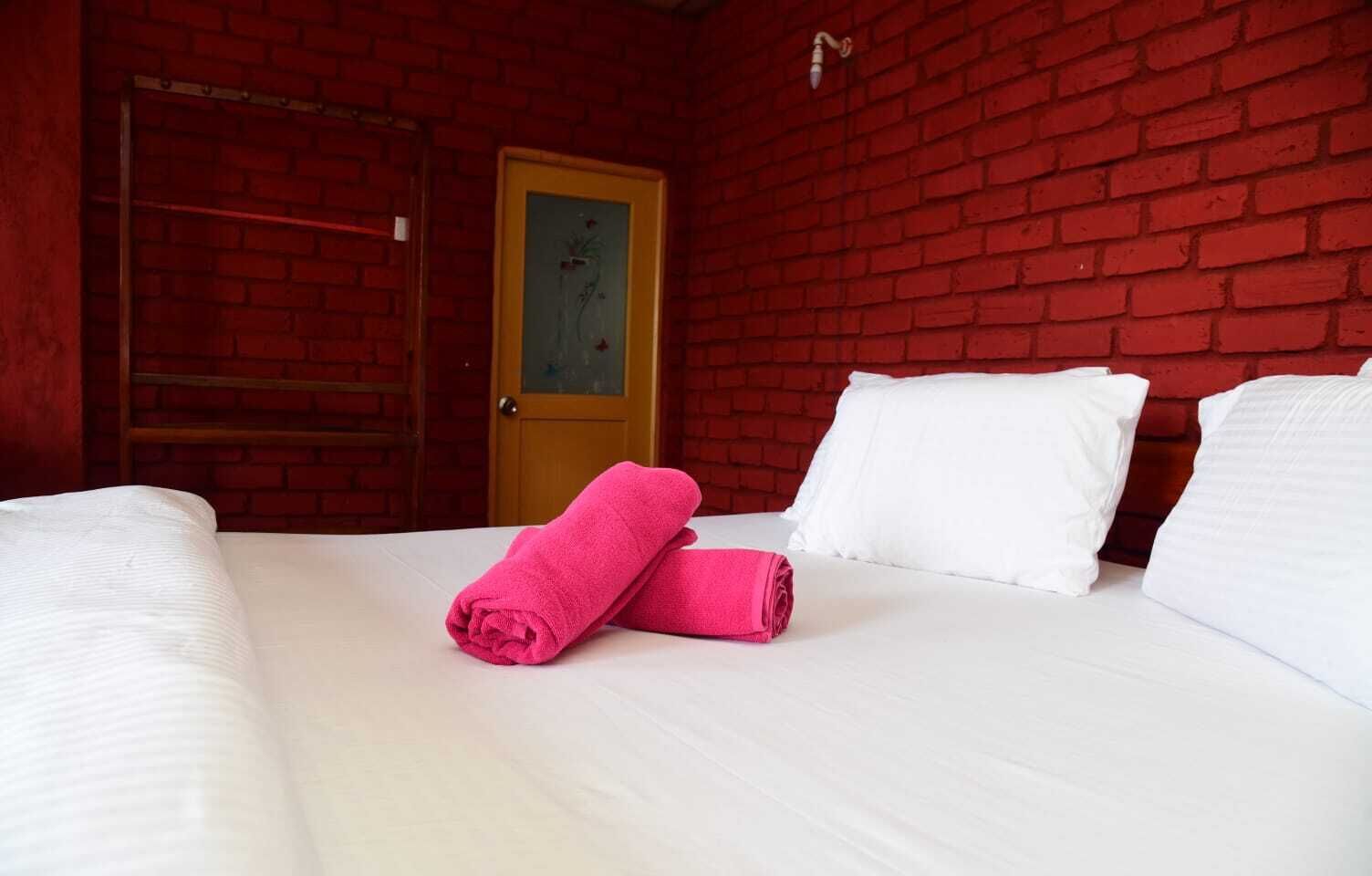 Habitación de hotel con pared de ladrillo rojo, puerta de madera y cama con ropa blanca y toallas rosas.