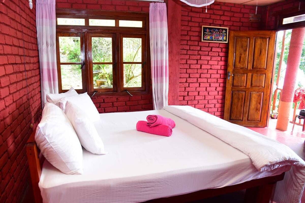 A bed with white linens, a pink towel, and a wooden door in a room with red brick walls.