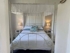 Una cama con dosel y cuatro columnas, con ropa de cama blanca, almohadones decorativos, dos lámparas de noche y un pie de cama con encaje.