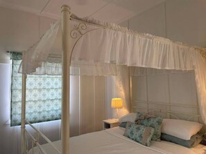 A four-poster canopy bed with a nightstand and lamp, a window with curtains, and a patterned wall.