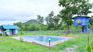 Uma piscina com estrutura de telhado azul, um pequeno edifício com fachada vermelha e azul, e uma estrutura elevada azul com detalhes amarelos.
