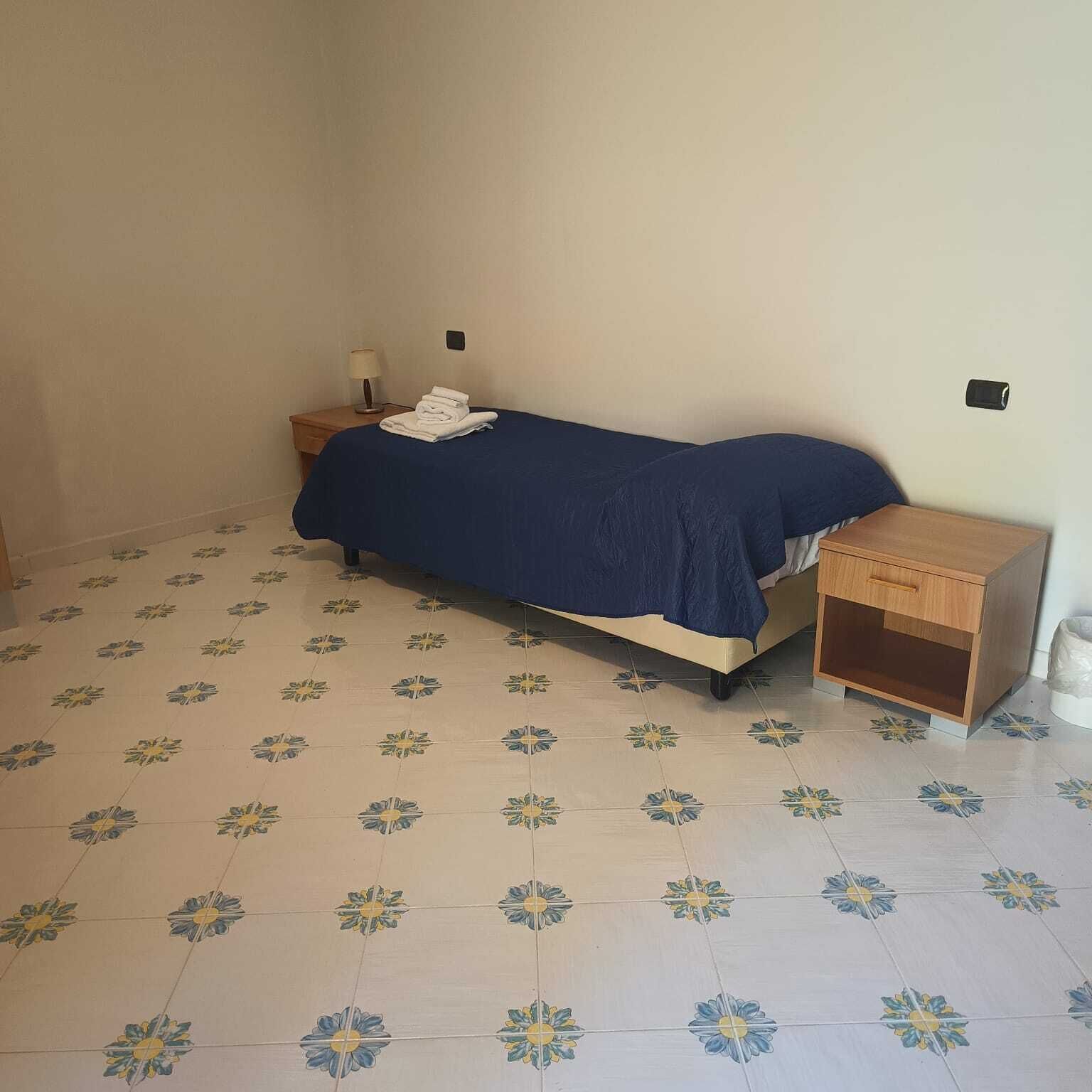 A single bed with a blue bedspread, a wooden nightstand with a lamp, and a wall-mounted switch. The floor has a floral tile pattern.