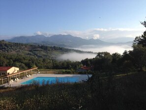 Zona de piscina con sillones de descanso, un porche cubierto y vistas a montañas y niebla.