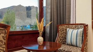 Una habitación con una mesa de madera, dos sillas estampadas, un florero con plantas secas y vistas a montañas a través de una ventana.